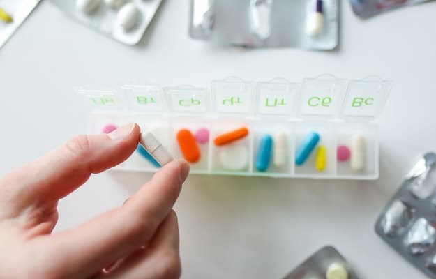 Pessoa organizando comprimidos em um porta-comprimidos semanal, com cartelas de medicamentos ao lado para facilitar o controle dos horários das doses.
