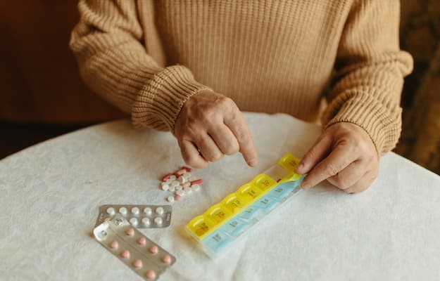 Pessoa organizando comprimidos em um porta-comprimidos semanal, com cartelas de medicamentos ao lado, para facilitar o controle dos horários das doses.