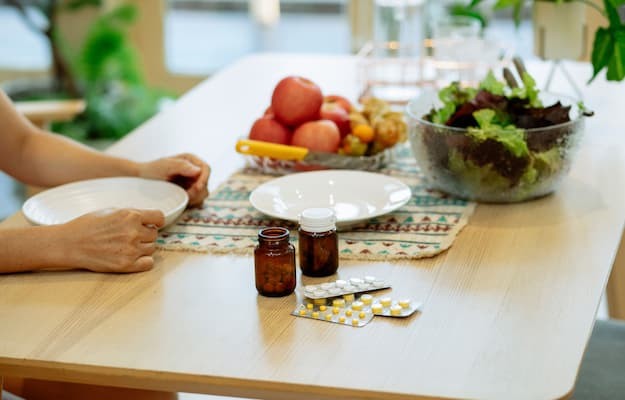 Medicamentos em frascos e cartelas sobre a mesa, ao lado de pratos, frutas e salada, e pessoa em dúvida se toma remédio antes ou depois de comer.