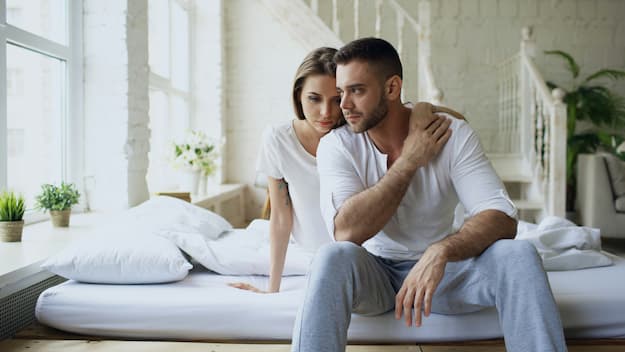 Casal sentado na cama com expressão de preocupação e distanciamento emocional com dificuldades relacionadas à disfunção sexual.