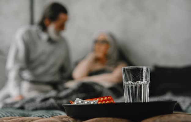 Bandeja com um copo de água e medicamentos em primeiro plano, enquanto um casal de idosos aparece desfocado ao fundo com a administração de remédios.