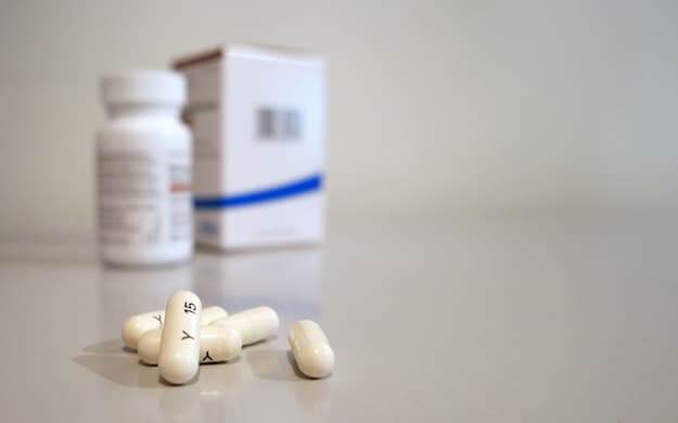 Frascos e caixa de medicamento ciclosporina, com cápsulas brancas espalhadas sobre uma superfície.